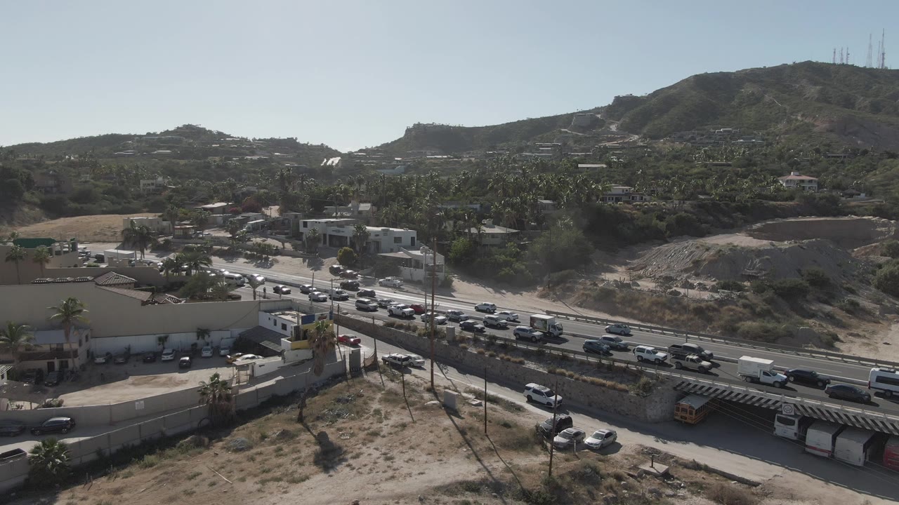 Opening frame of traffic moving along a multi-lane road in Costa Azul Los Cabos, with dry foreground scrub, low buildings, hillside terrain, and no visible people.