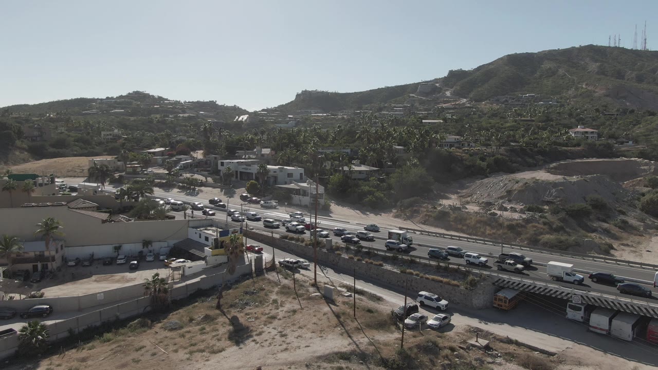 Traffic continues through Costa Azul Los Cabos, with vehicles in both directions, roadside walls and buildings in the midground, hills beyond, and no visible people.