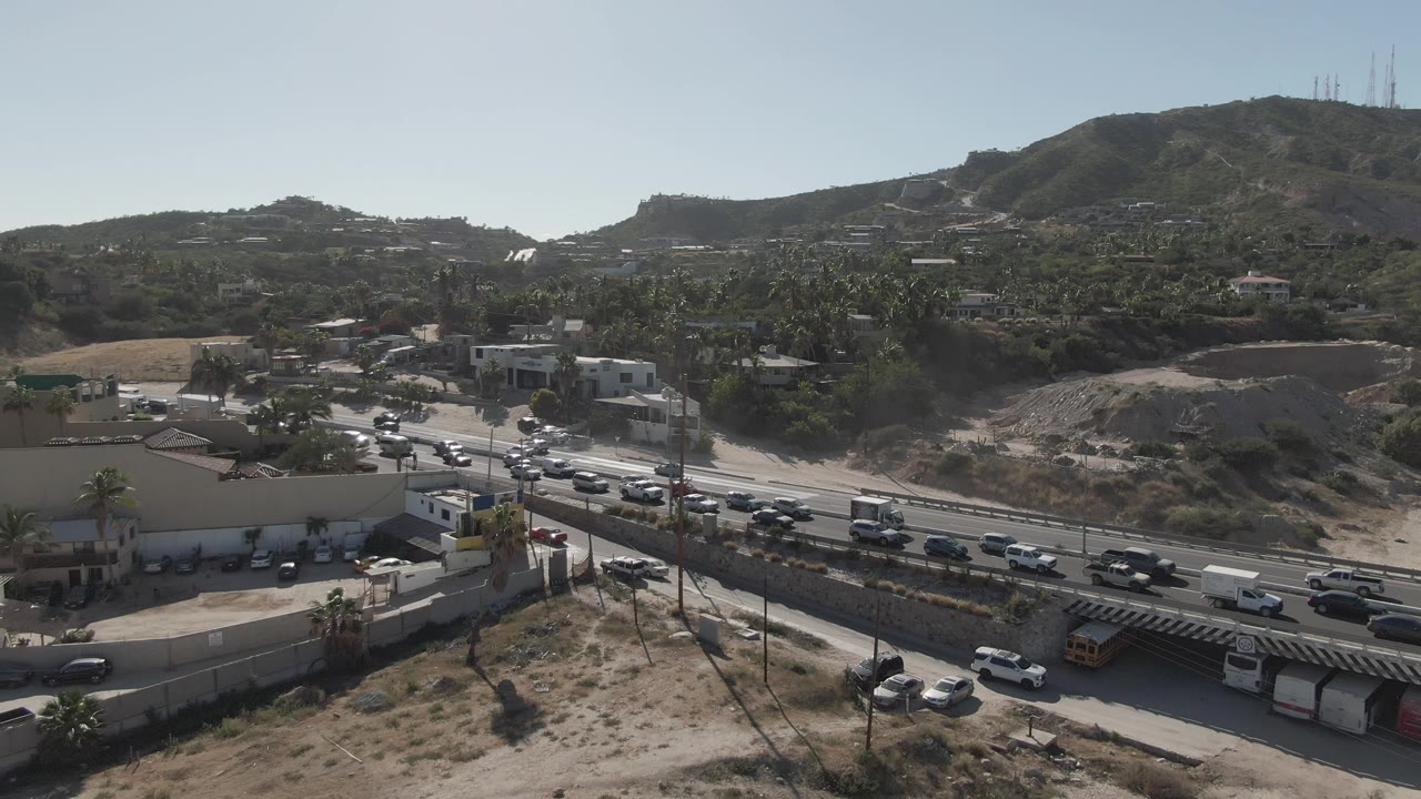 A wide frame shows steady vehicle flow on the Costa Azul Los Cabos road, with dry terrain in front, clustered buildings near the lanes, distant hills, and no visible people.