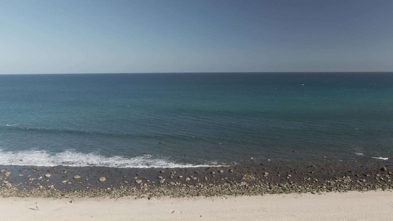 Aerial frame at Costa Azul Los Cabos showing pale sand, a dark rocky shore band, blue-green ocean water, small white wave foam, and no visible people.