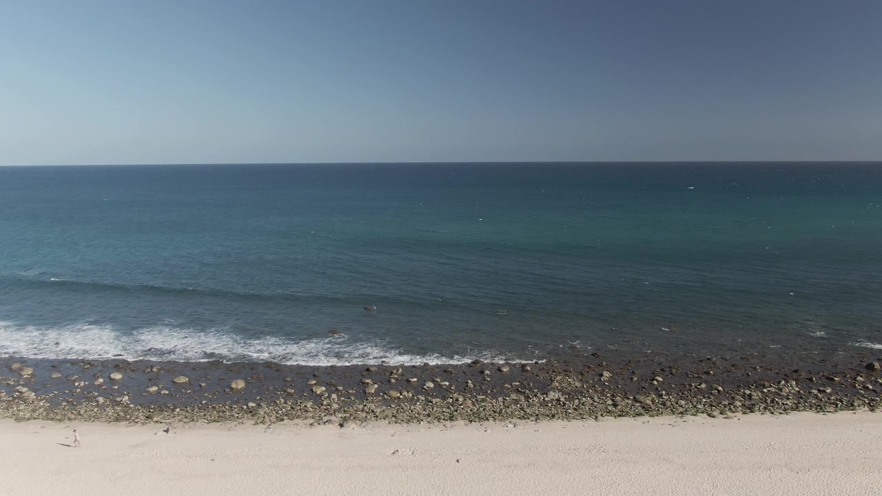 Small waves break along a rocky strip beside a sandy beach.