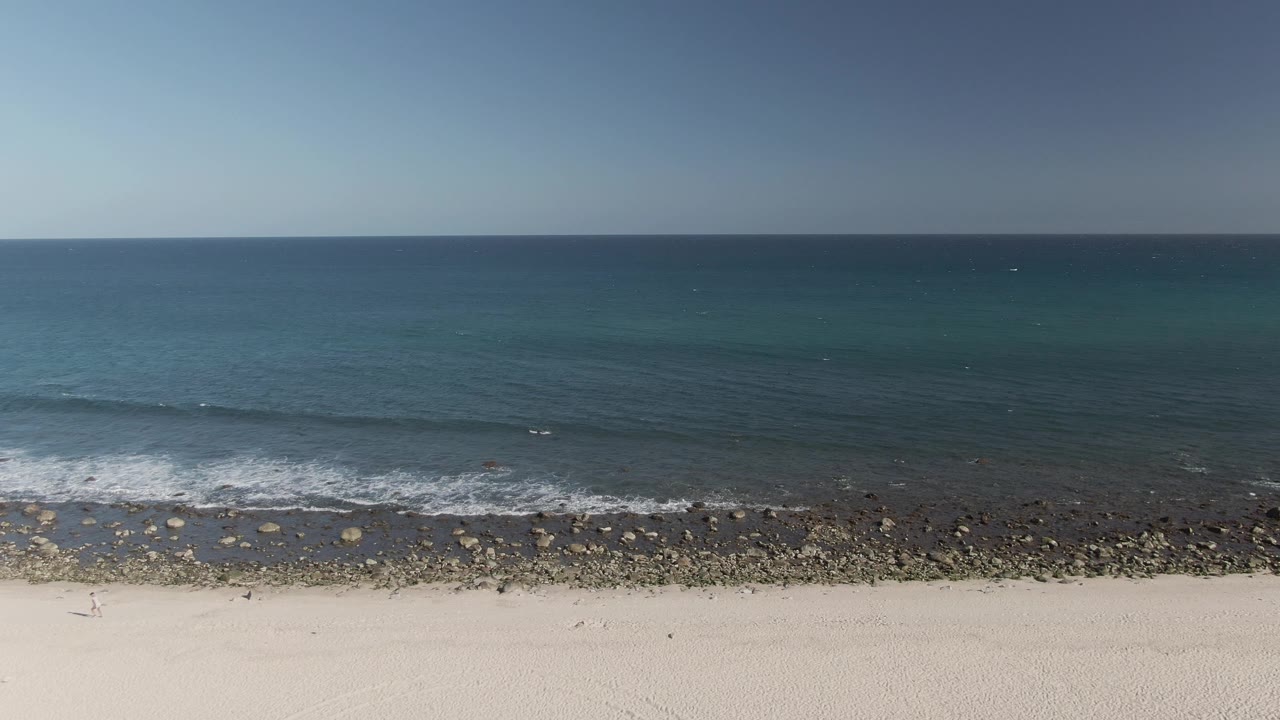 Aerial view at Costa Azul Los Cabos with scattered rocks along the waterline, a narrow foam band, open blue sea, pale foreground sand, and no visible people.