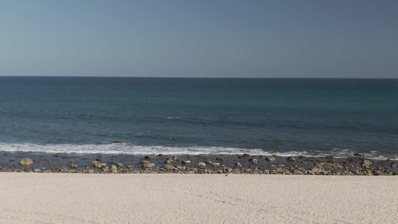 Wide view of Costa Azul Los Cabos beach with white sand in front, a rocky shoreline, small ocean waves, clear blue sky, and no visible people.