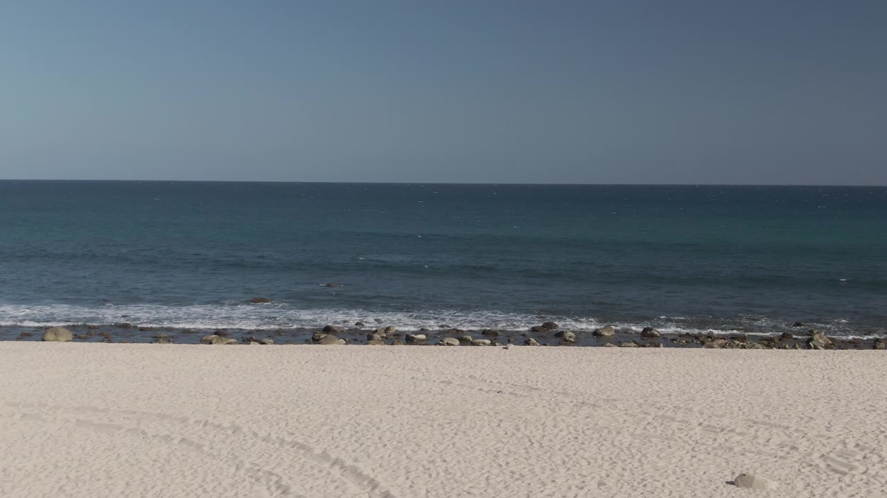The Costa Azul Los Cabos shoreline fills the frame with empty white sand below, rocks along the surf, a level ocean horizon, and no visible people.