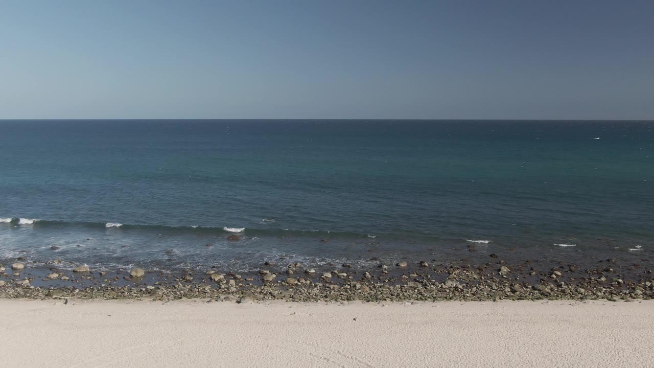 At Costa Azul Los Cabos, blue ocean stretches to a level horizon while small waves break across a rocky shoreline above pale sand, with no visible people.