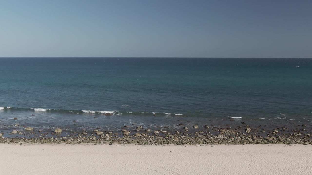 Small white waves approach a rocky shore beneath a clear blue sky.