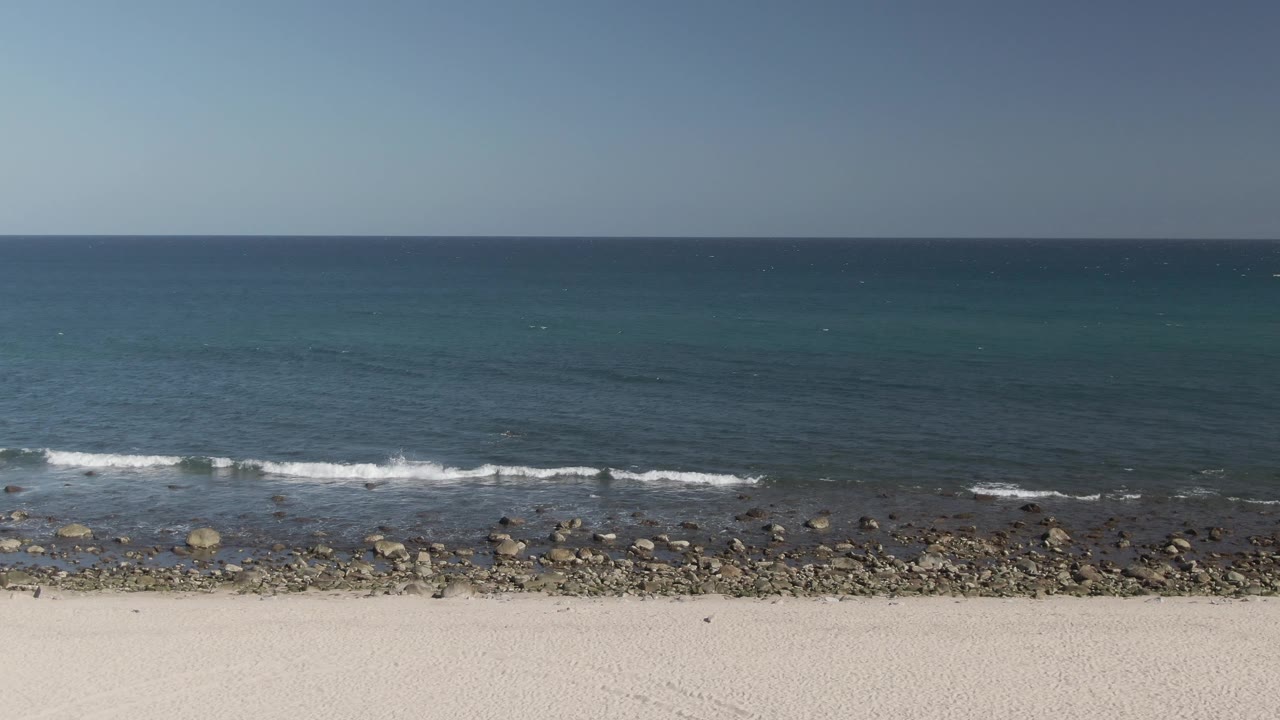 At Costa Azul Los Cabos, darker blue water sits beyond a broken band of rocks while low waves leave white foam near the shore and no visible people appear.