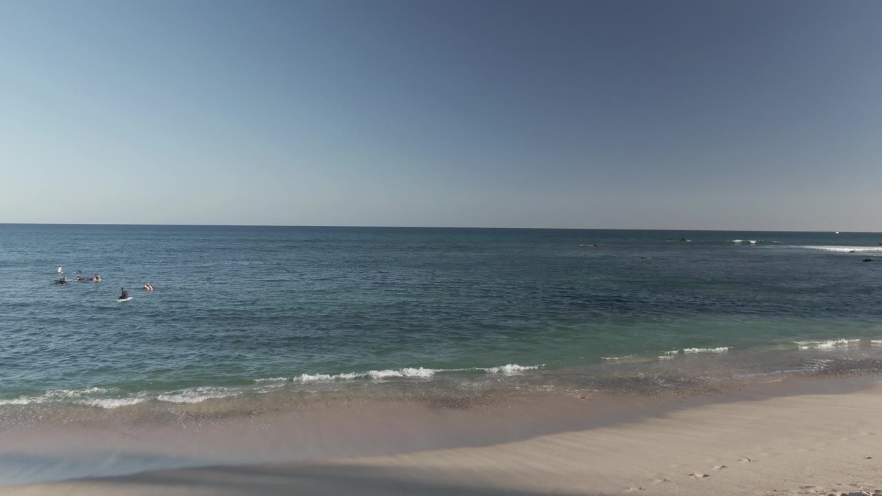 Wide elevated view of Costa Azul Los Cabos beach with sandy shore, small waves, blue-green water, swimmers offshore, and clear sky.