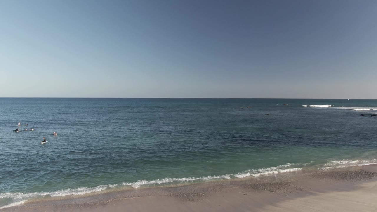 Costa Azul Los Cabos beach view with a thin line of surf, darker offshore water, scattered swimmers to the right, and bright sky above the horizon.
