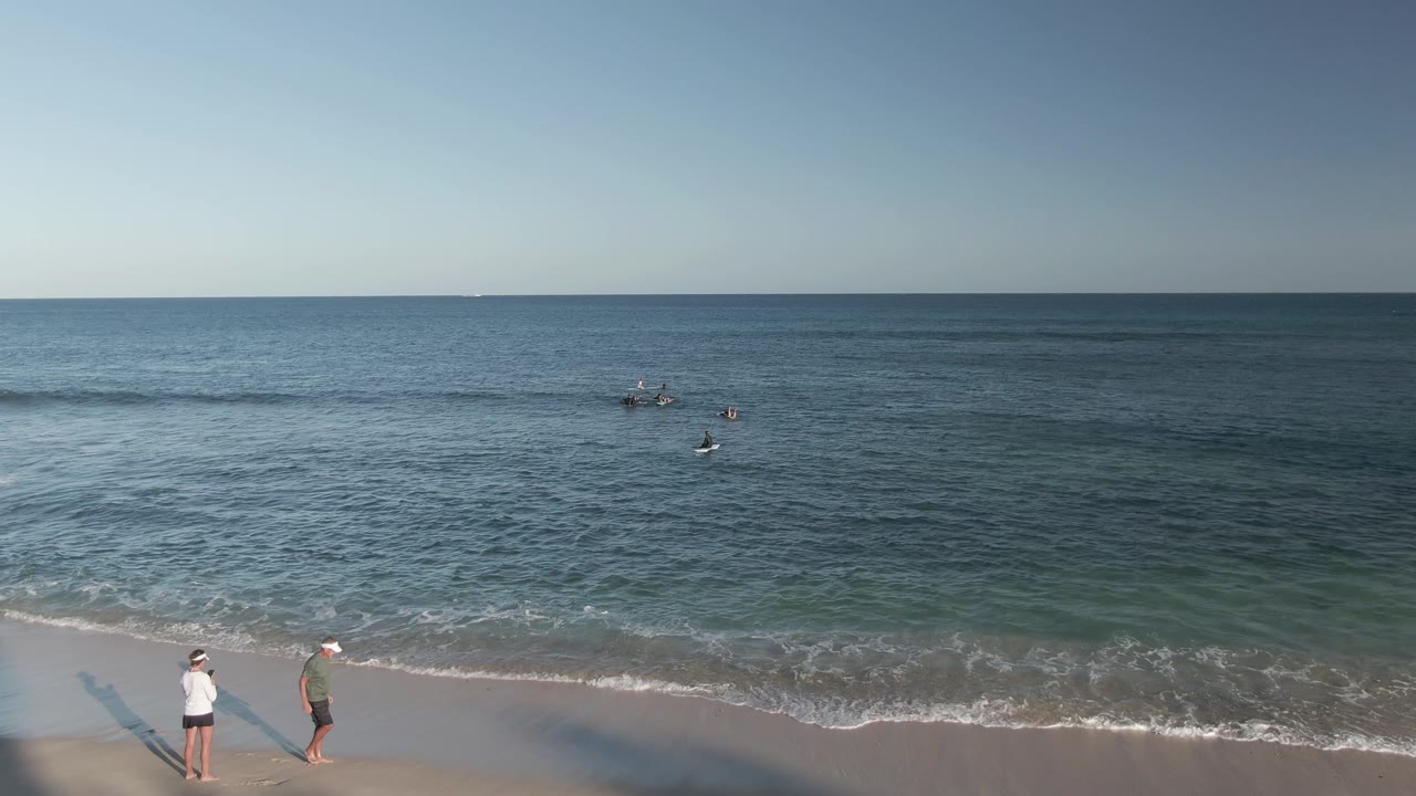 Costa Azul Los Cabos beach at 0:00 with two adults near the lower left edge, scattered swimmers offshore, and small waves reaching the wet sand.