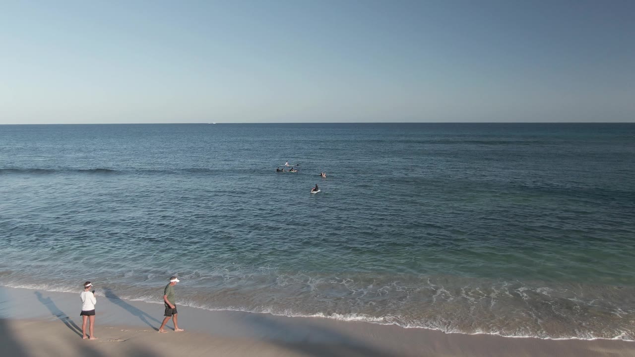 Two beach walkers pass wet sand while swimmers remain offshore.