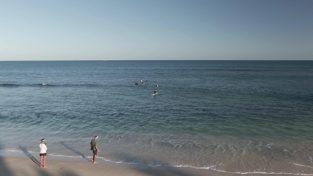 Costa Azul Los Cabos beach at 0:02 with a white-shirted walker near lower center-left, a darker-clothed walker near lower center, and multiple swimmers floating beyond the shore break.