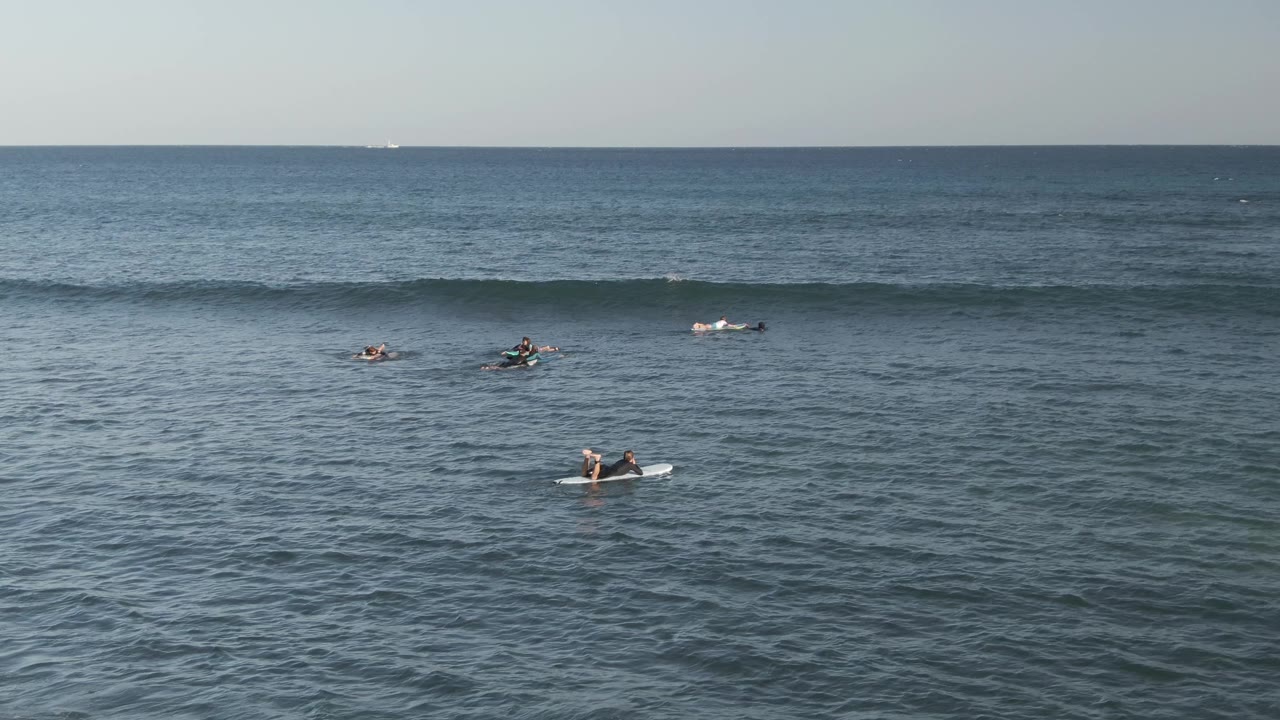 At Costa Azul Los Cabos, distant surfers sit on boards across blue ocean water as a long wave line starts to rise near the horizon.
