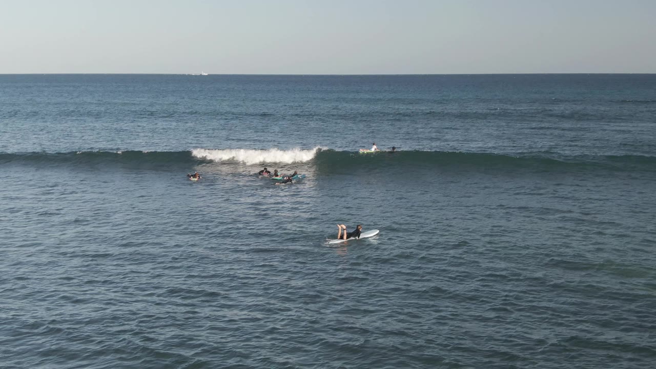 The wave breaks harder on the right side of the frame while scattered surfers remain on boards in the blue-green ocean at Costa Azul Los Cabos.