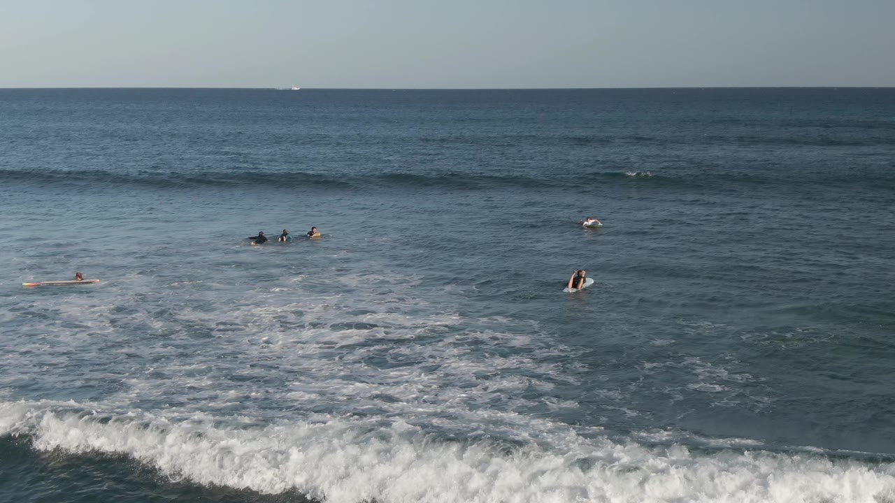 At Costa Azul Los Cabos, surfers sit and paddle on boards across blue water while a foamy wave edge enters the lower foreground.