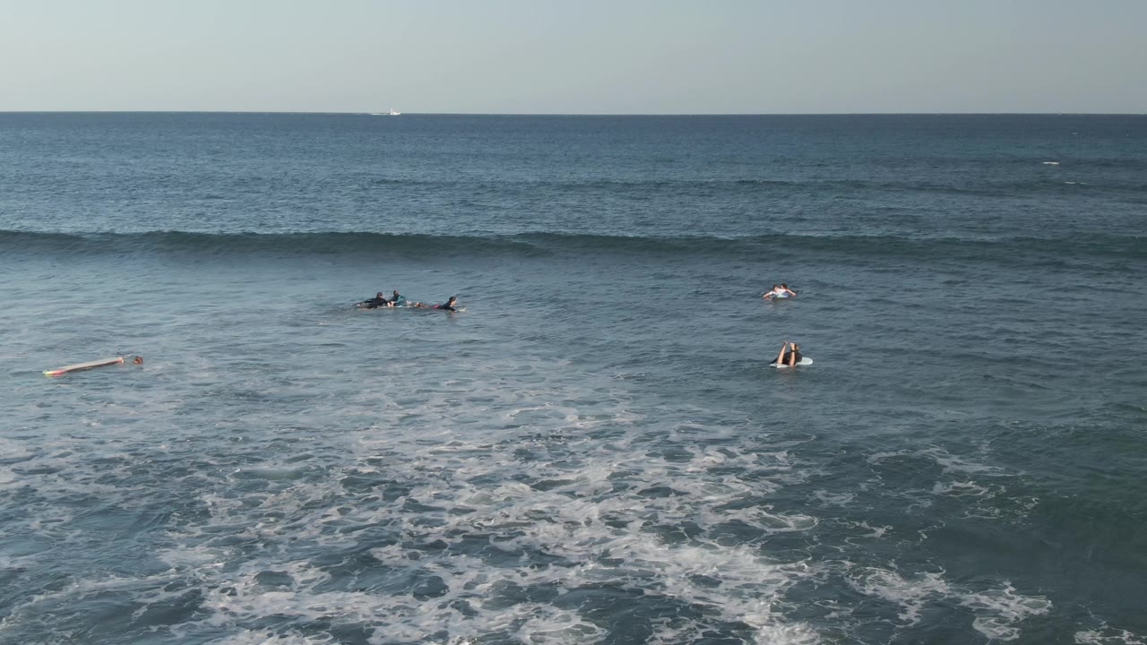 At Costa Azul Los Cabos, scattered surfers and boards sit in the midground while foam patterns spread below and a broad wave rolls across the back of frame.