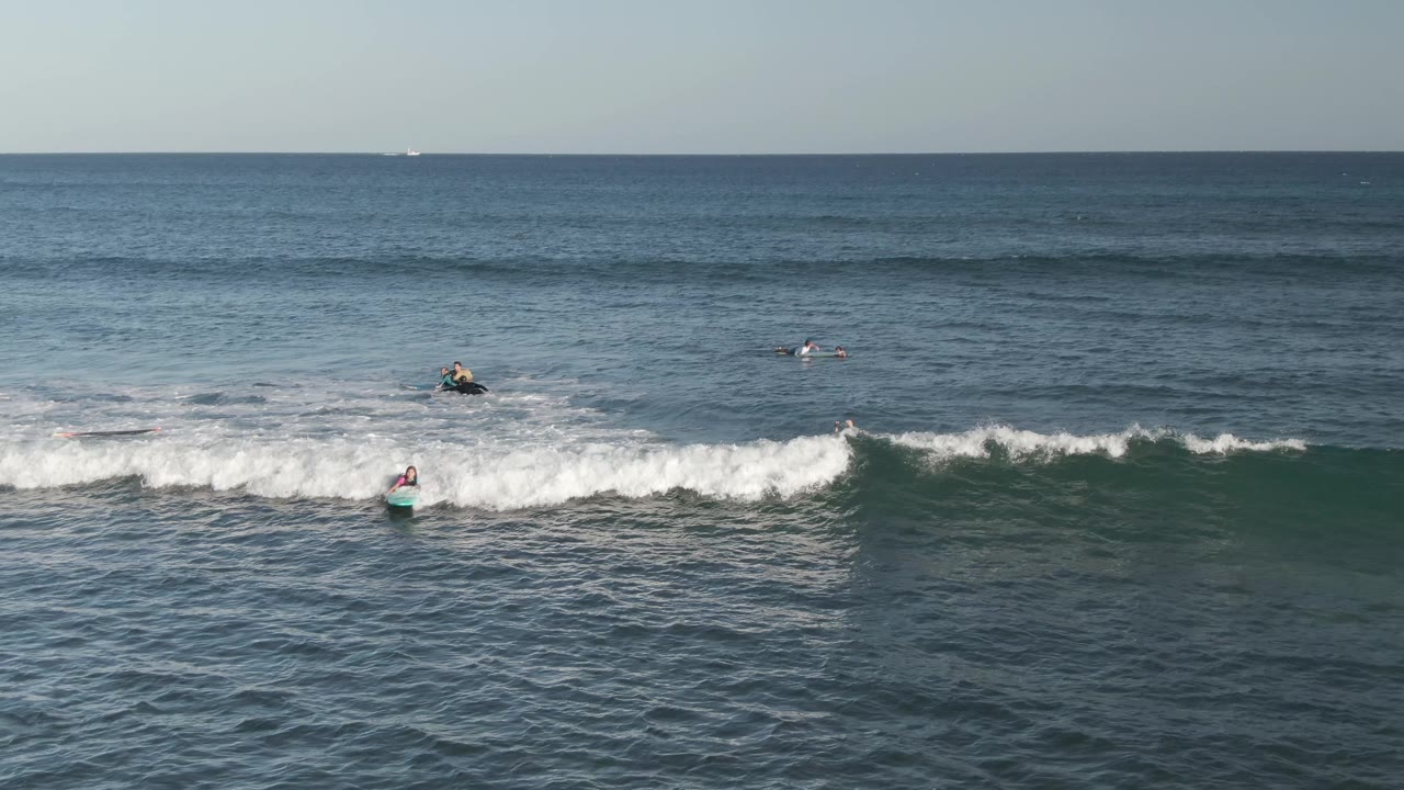 A surfer rides down the face of a breaking wave at Costa Azul Los Cabos while scattered surfers and bodyboarders wait in the surrounding blue water.
