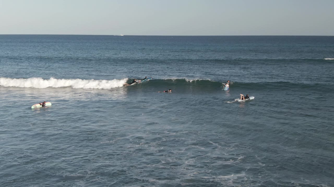 Surfers and bodyboarders are spaced across the blue ocean at Costa Azul Los Cabos as a white wave begins to break near the left side of the frame.
