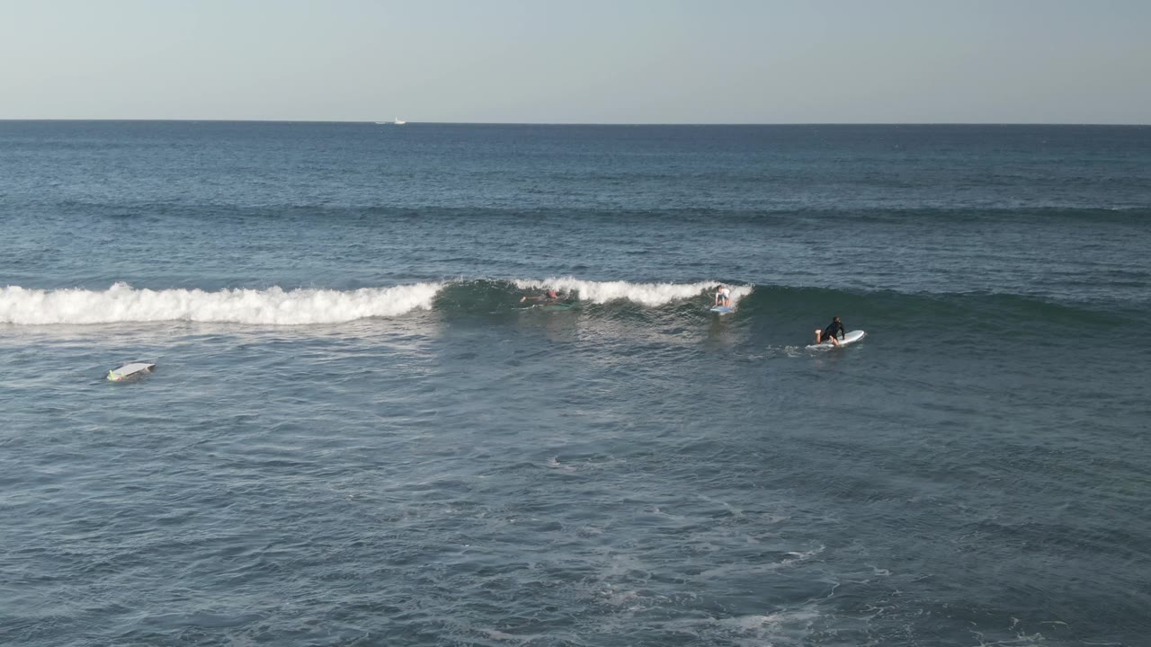 A low white wave breaks across the midground at Costa Azul Los Cabos while several surfers paddle nearby in deep blue water.