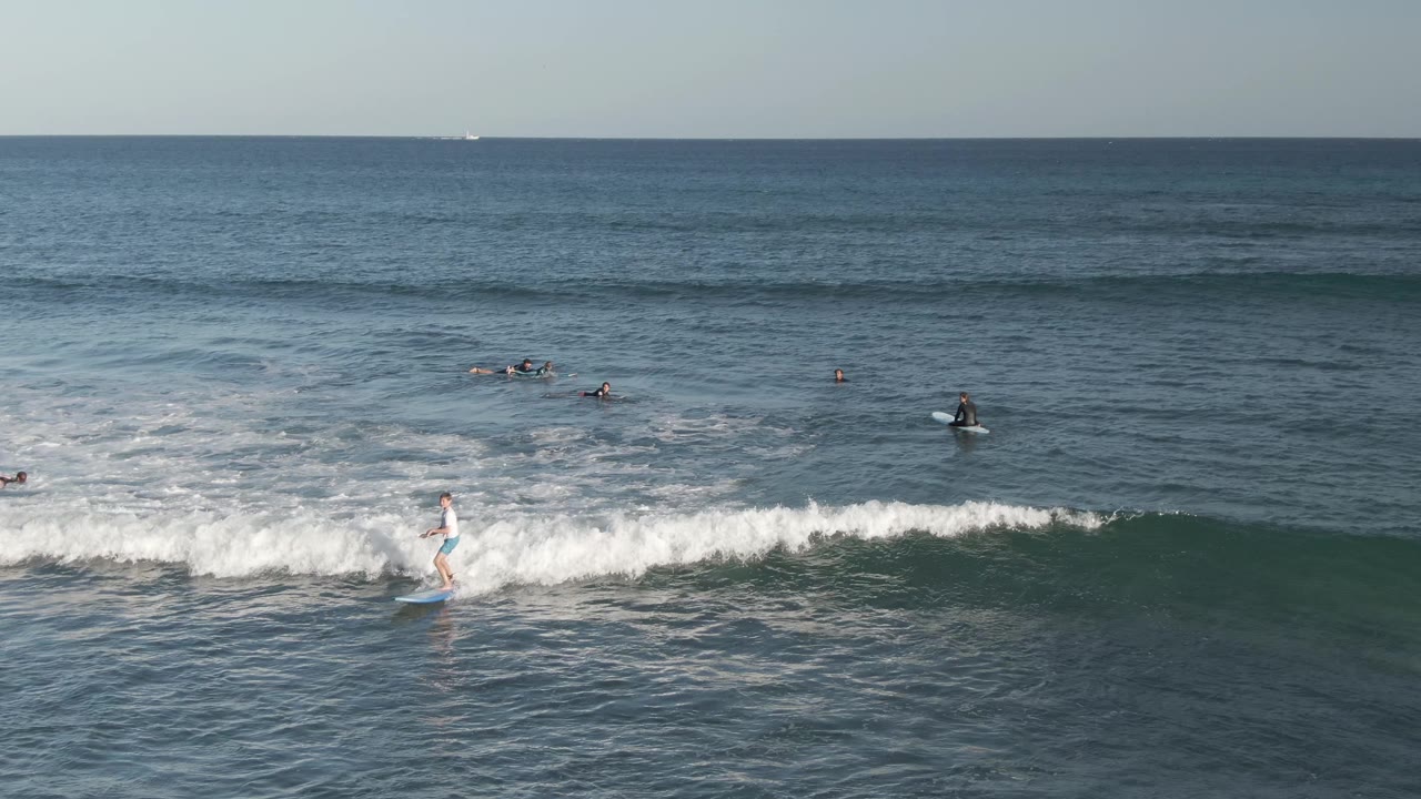 At Costa Azul Los Cabos, a surfer in light gear rides a small breaking wave while several other surfers sit farther out under a flat ocean horizon.