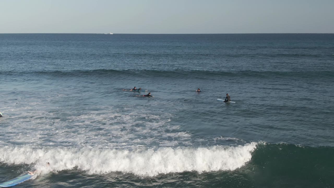 At Costa Azul Los Cabos, the wave throws more whitewater across the foreground and a light blue surfboard is visible near the breaking section while surfers remain beyond it.