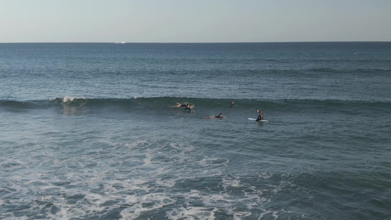Wide aerial view at Costa Azul Los Cabos with small surfers spread across blue-green water, foamy surf in the foreground, and a straight ocean horizon behind them.