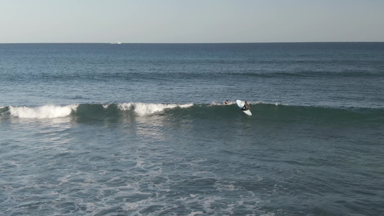 Aerial view at Costa Azul Los Cabos showing surfers scattered along a breaking wave, with white spray on the wave face and calmer open sea beyond.