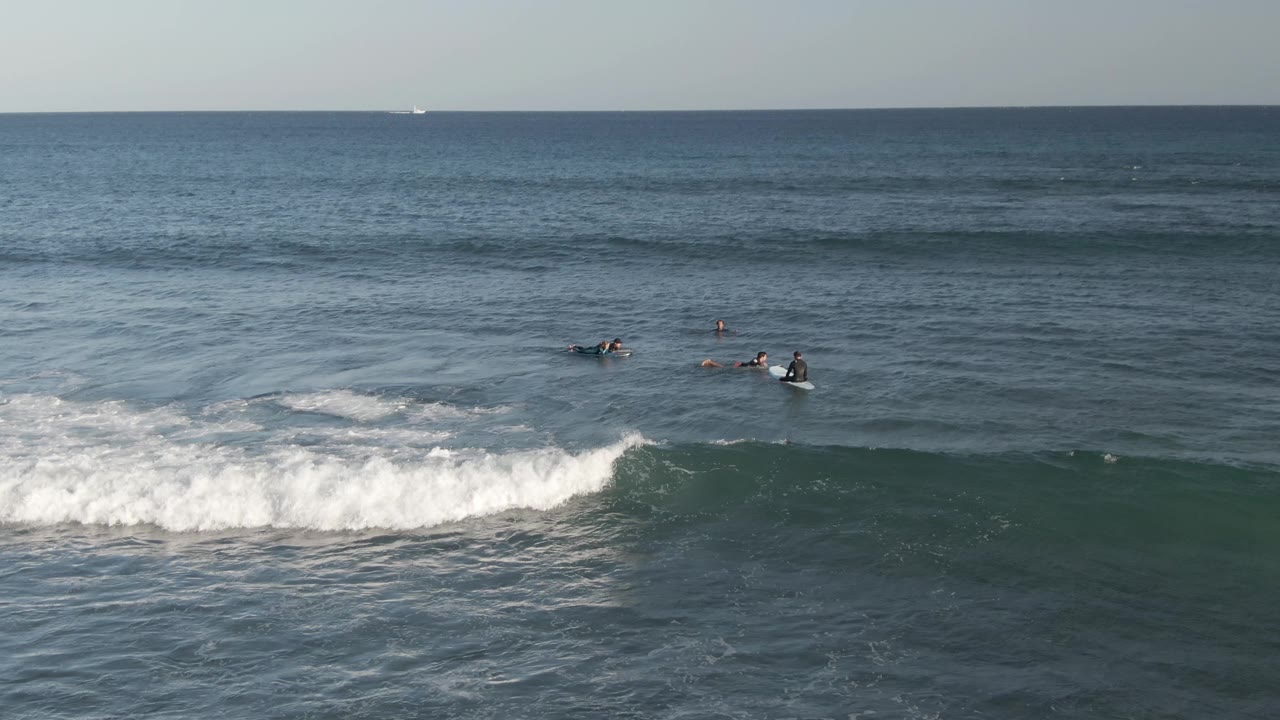 Small surfers on boards paddle beyond whitewater at Costa Azul Los Cabos, with a breaking wave in the foreground and open blue water to the horizon.