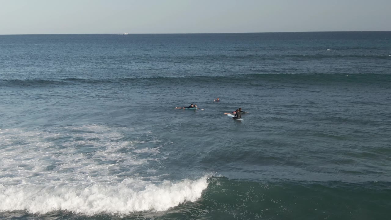 A cluster of surfers is spread across the midground at Costa Azul Los Cabos as a thicker white foam line breaks across the lower right of the frame.