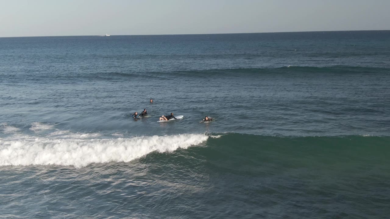 At Costa Azul Los Cabos, several surfers sit or paddle on boards beyond a band of breaking whitewater, with a broad blue ocean and straight horizon behind them.