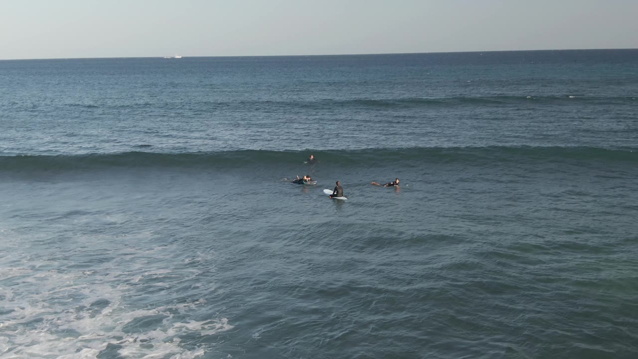 At Costa Azul Los Cabos, several surfers are spread across blue-green water while a long wave begins to rise near the horizon side of the frame.