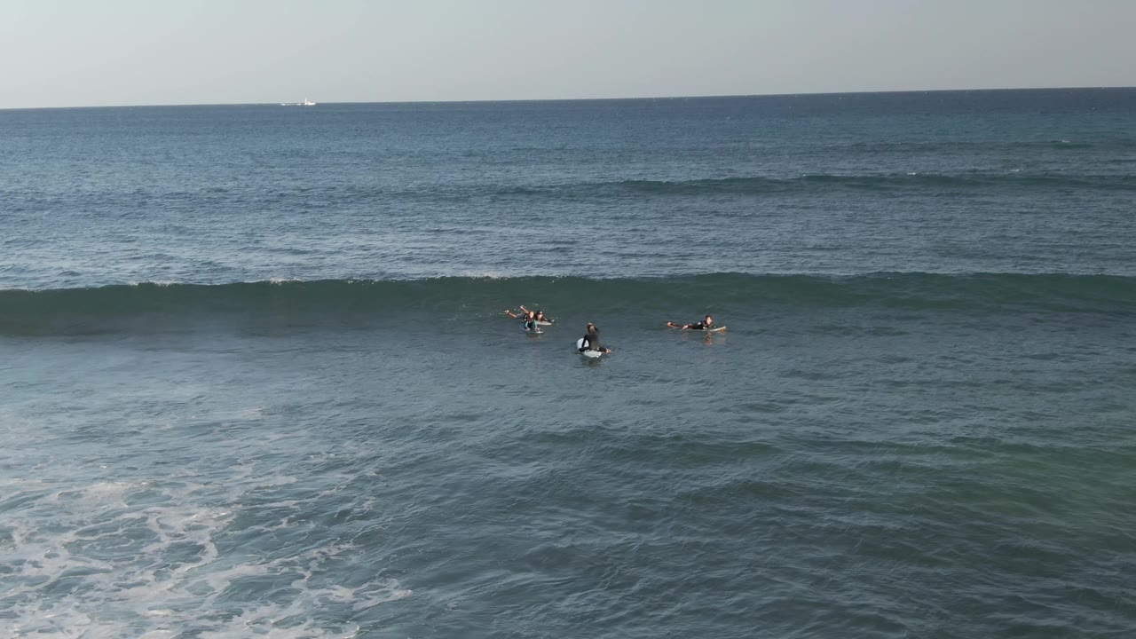 A surfer sits in the center as a wave starts to crest on the right.