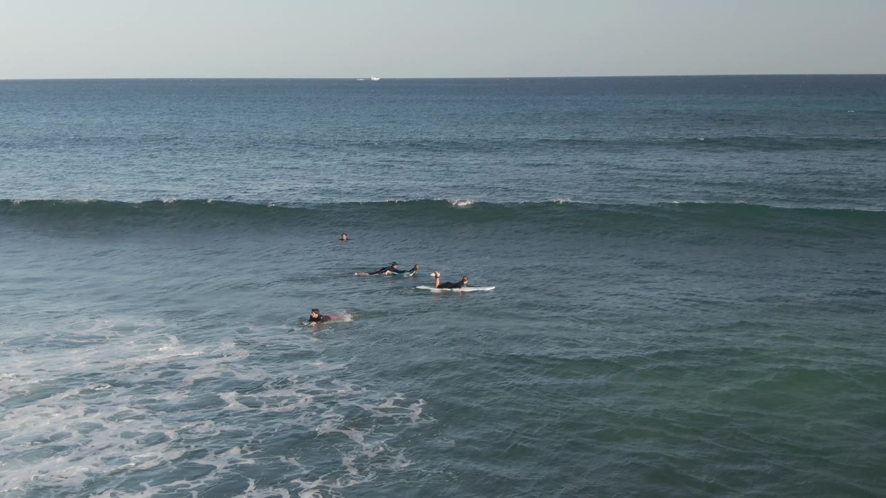 Surfers paddle on boards in the blue-green ocean at Costa Azul Los Cabos, with white foam in the lower frame and a long wave forming behind them.