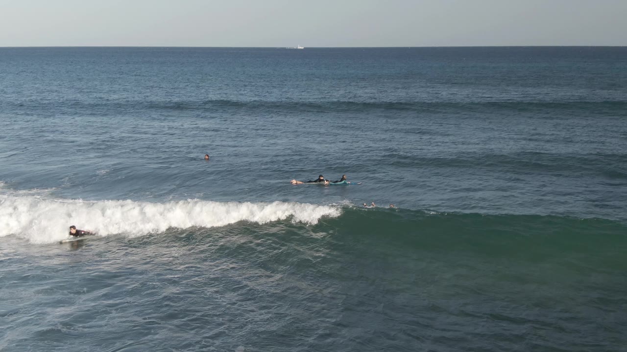 Distant surfers paddle near a breaking wave in Costa Azul Los Cabos, with blue-green ocean, white foam, and a level horizon.