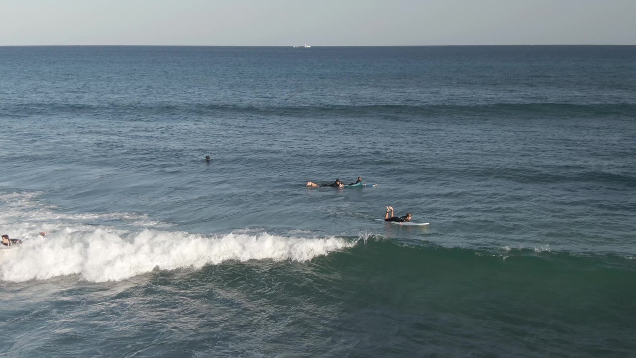 Several distant surfers and bodyboarders sit or paddle beyond whitewater in Costa Azul Los Cabos, with open blue water stretching to the horizon.