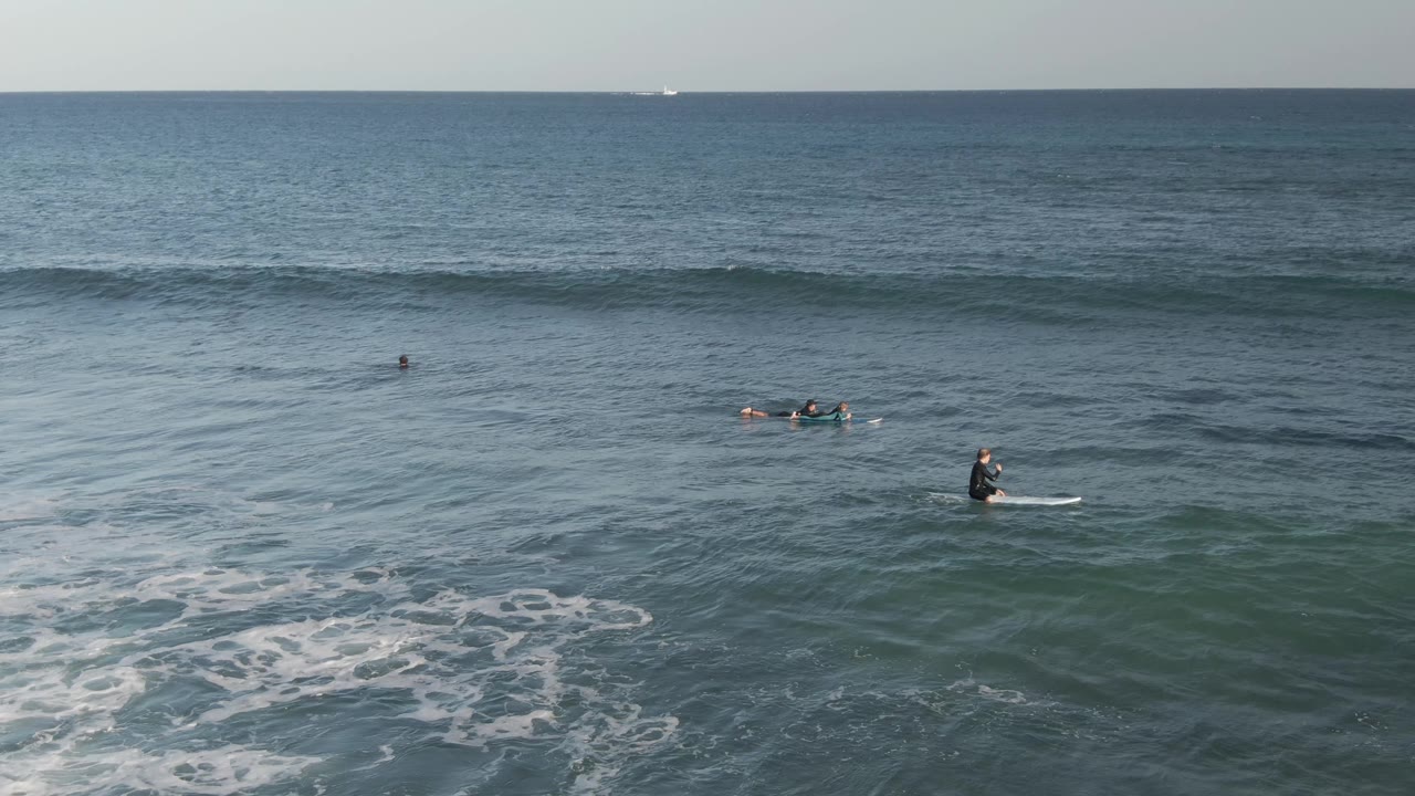 Surfers and swimmers sit spread across the blue-green water at Costa Azul Los Cabos as a long wave line forms beneath a level ocean horizon.