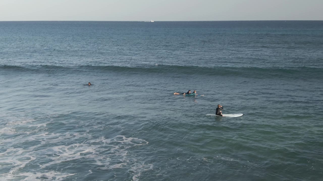 Several surfers wait on boards as a wave lifts behind them.