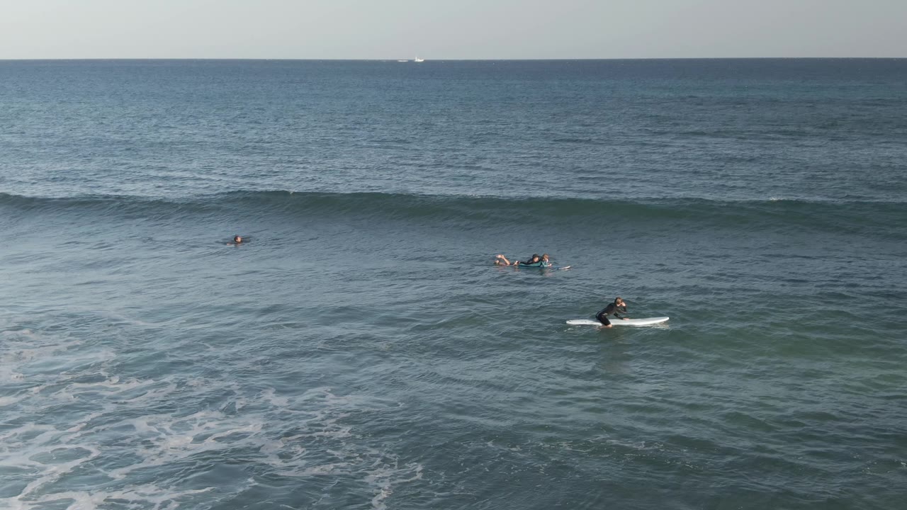 A wide Costa Azul Los Cabos ocean frame shows small surfers on boards below a smooth, unbroken wave line and a calm open-water horizon.