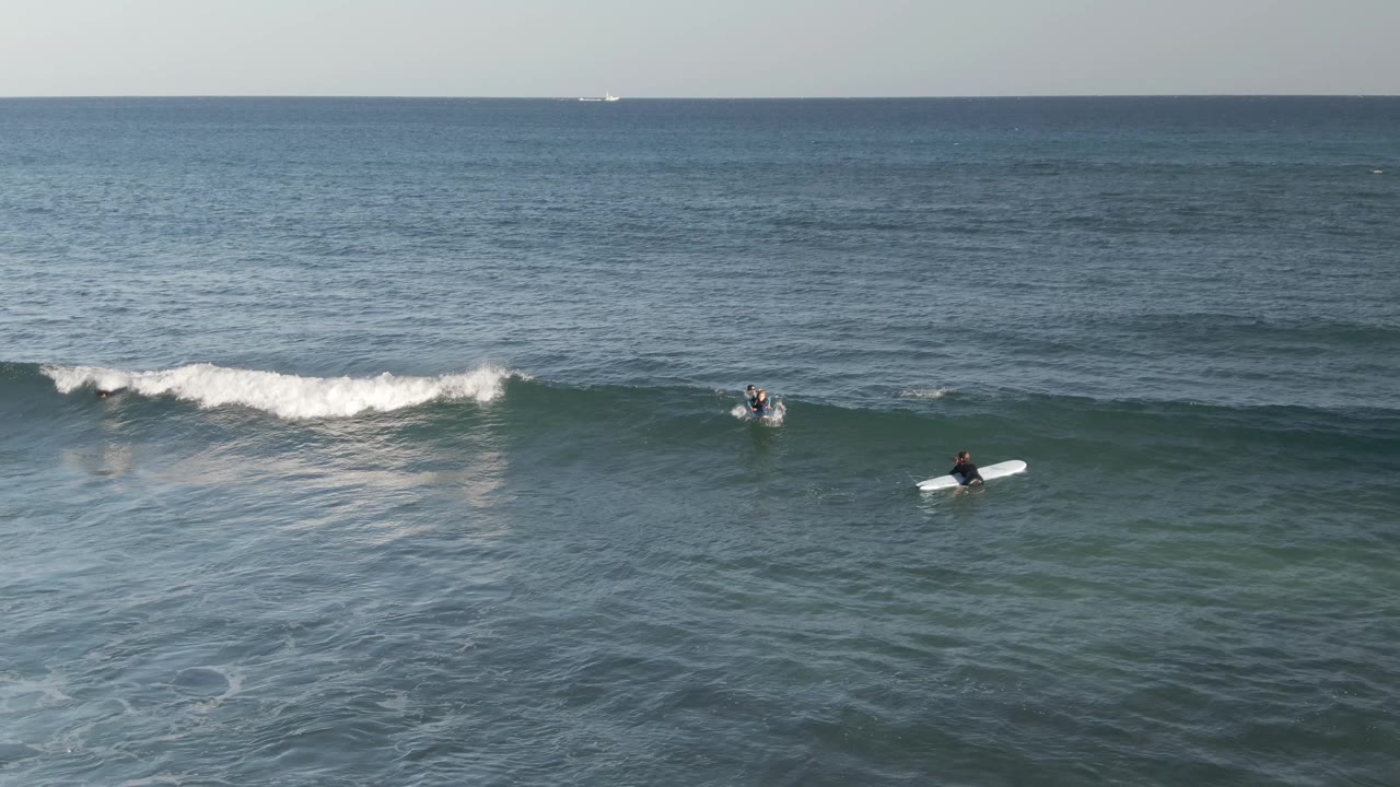 Aerial view at Costa Azul Los Cabos of several surfers spaced across blue-green water as a low white wave begins to break near the left side of frame.