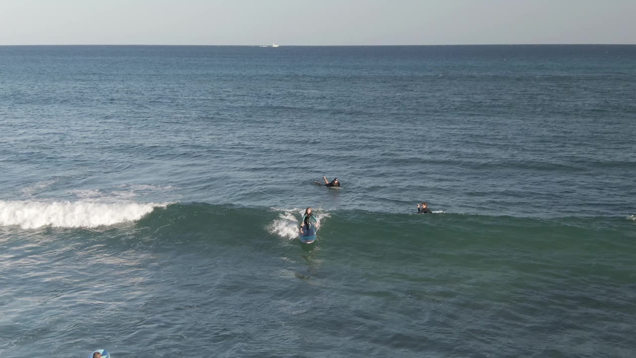 Aerial view at Costa Azul Los Cabos showing one surfer starting down the face of a breaking wave, with other surfers beyond the foam line in open water.