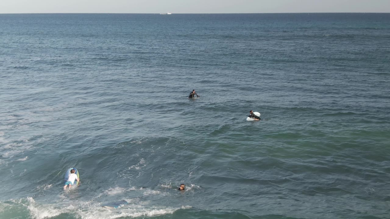 Aerial view of surfers spread across blue-green ocean water at Costa Azul Los Cabos, with white foam near the lower edge and a flat horizon in the distance.
