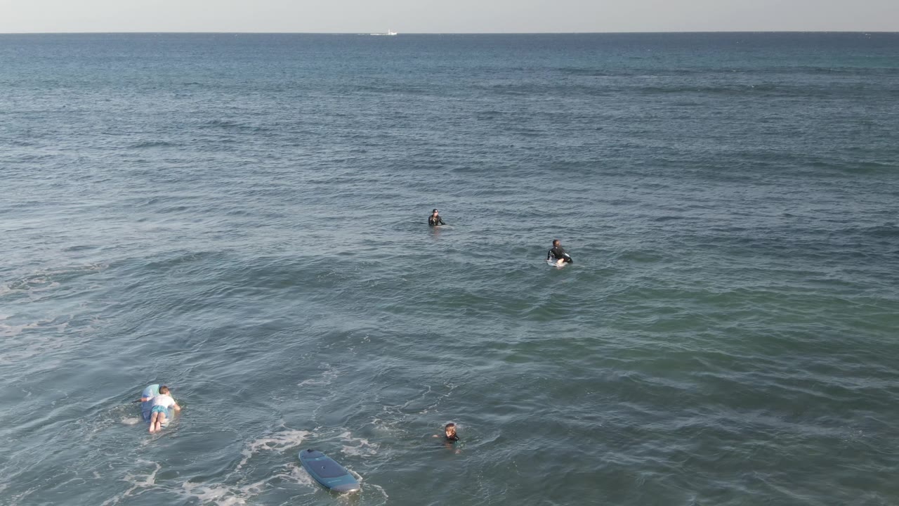 Aerial view at Costa Azul Los Cabos with surfers sitting and paddling in blue-green water, a surfboard partly visible near the bottom edge, and thin foam lines on the swell.