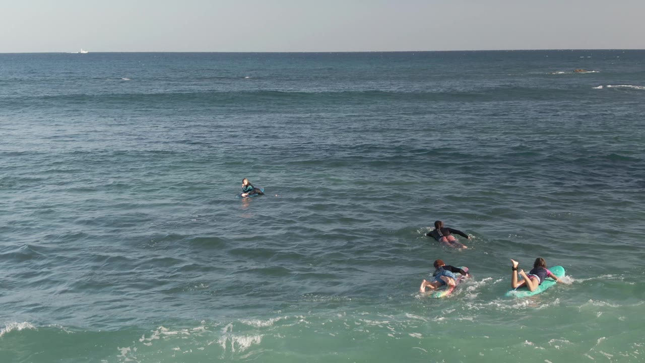 At Costa Azul Los Cabos, several people paddle on surfboards and bodyboards in blue-green water, with small waves and a level horizon behind them.