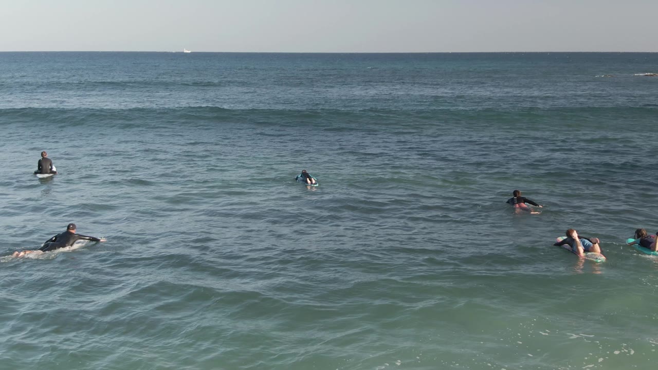 Surfers float and paddle on boards in blue green water at Costa Azul Los Cabos, with a low swell line forming behind them near the horizon.
