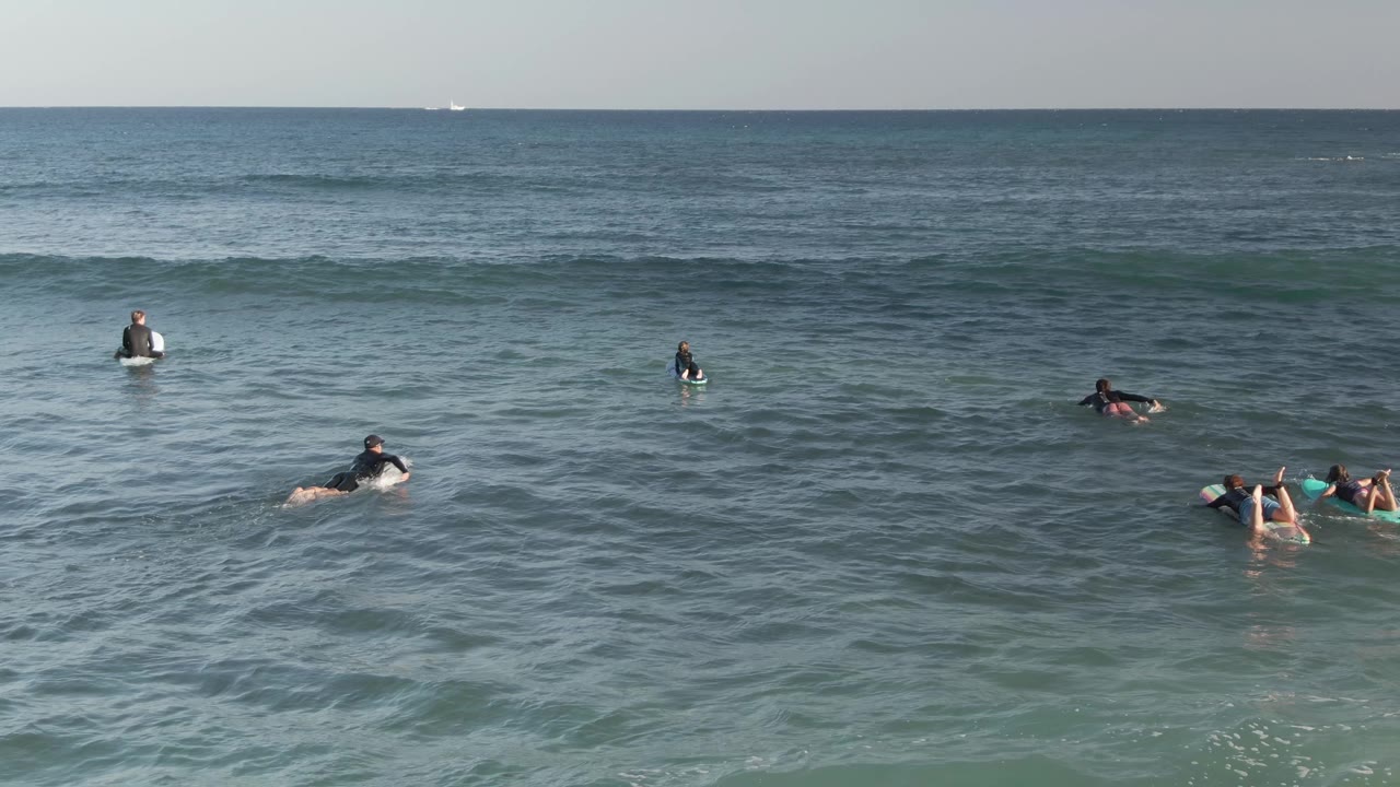 Several surfers paddle or sit on boards at Costa Azul Los Cabos, with one low wave band stretching across the background water.
