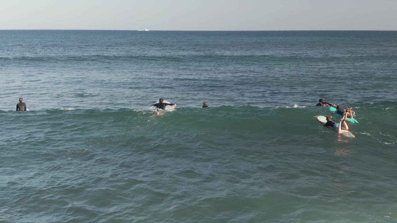 Wide elevated view of surfers waiting on boards in blue-green ocean water at Costa Azul Los Cabos, with a low swell forming near the horizon line.