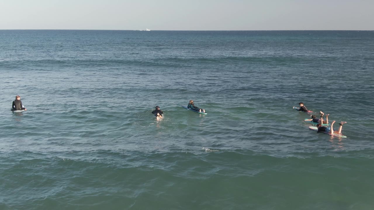 A broad surf lineup at Costa Azul Los Cabos is visible from above, with riders spaced across rolling ocean texture and a small boat far on the horizon.
