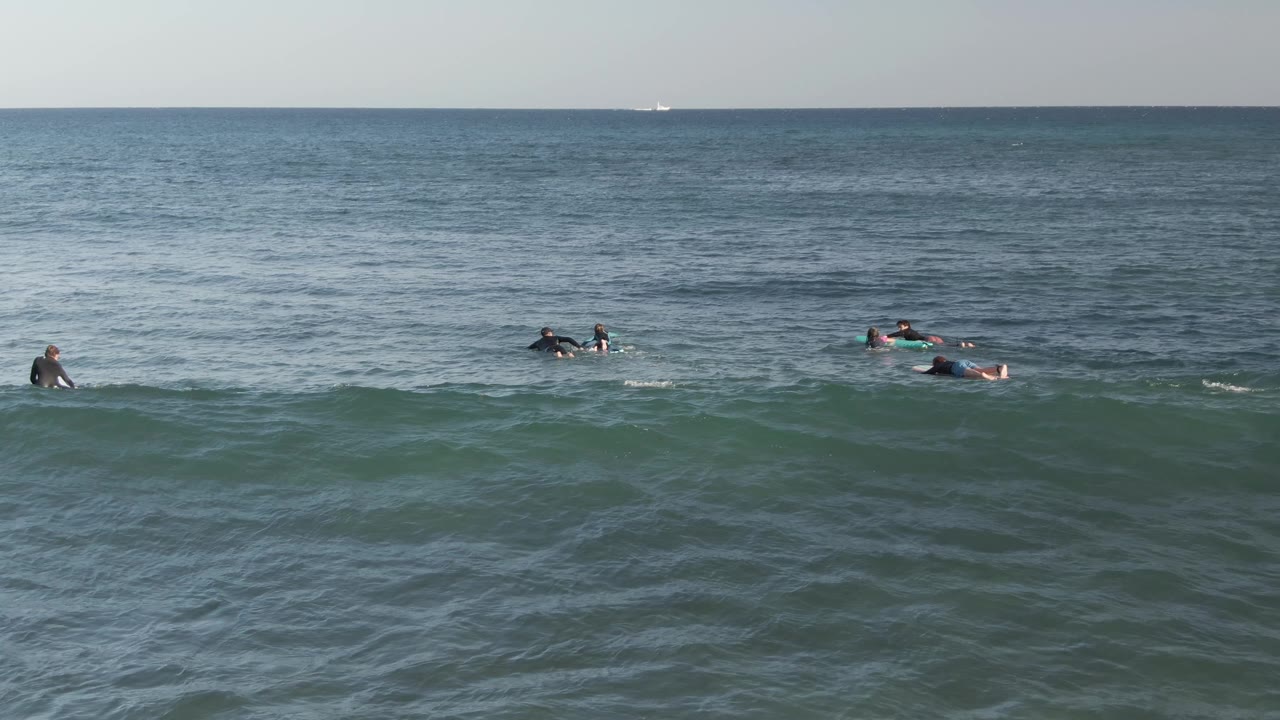 Several surfers in wetsuits sit and paddle on surfboards in blue-gray water at Costa Azul Los Cabos, with a small wave forming in the foreground.