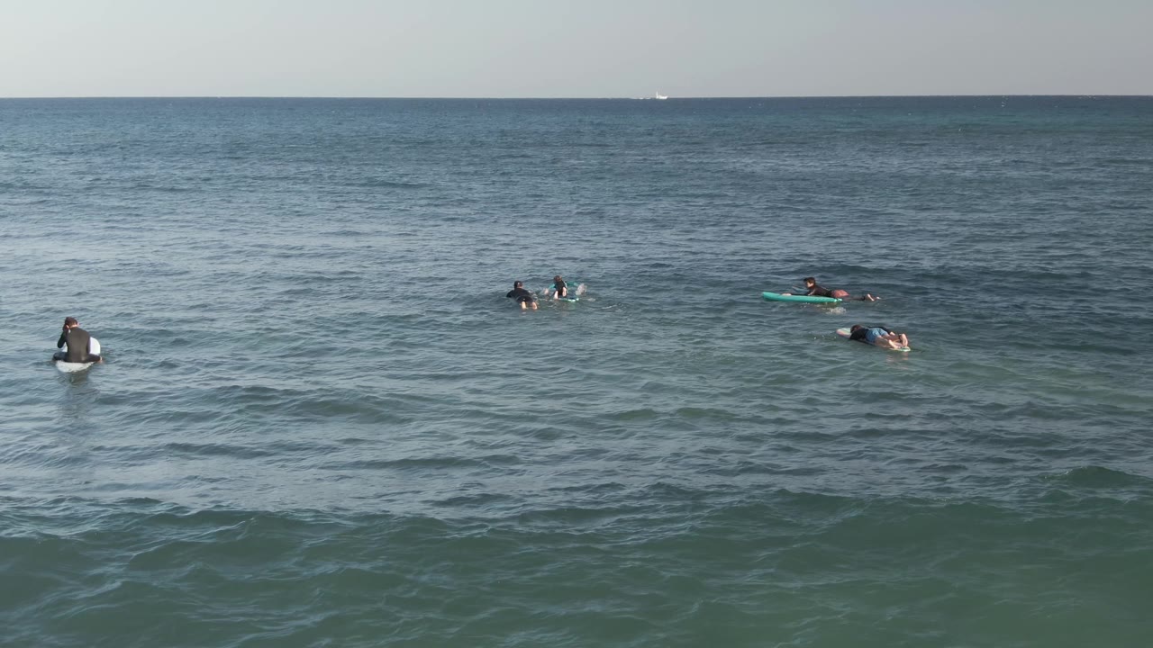 A central surfer sits upright on a board at Costa Azul Los Cabos while other surfers paddle nearby across textured blue-gray water.