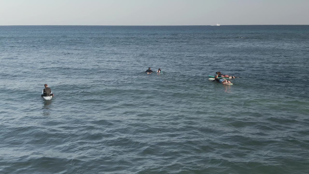 Surfers are spaced across the center of the frame at Costa Azul Los Cabos, with a seated board rider nearer the foreground and faint vessels on the distant horizon.